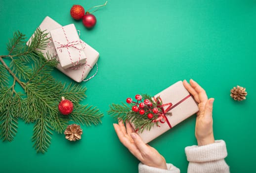 Christmas or New Year celebration green paper festive background with female hands holding wrapped gift box, decoration fir tree, cones, berries, sparkly red balls. Giving presents concept..