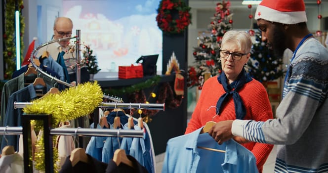 Worker assisting senior customer with picking right outfit to give as present to husband for upcoming xmas holiday season. Client browsing through clothes in festive ornate fashion shop