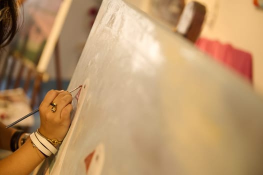 Details on female artist's hands working on painting on canvas, in creative art studio, holding a palette with oil paints and brush. Creative hobby and occupation. Fine art class. Close-up