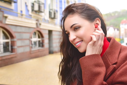 A smiling charming lady listens to music on the street using wireless headphones.
