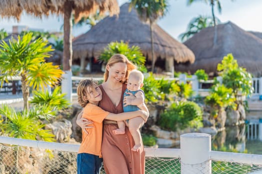 In a tropical paradise, a mom lovingly embraces her baby and 10-year-old son amid lush palm trees and thatched roofs, creating heartwarming family memories.
