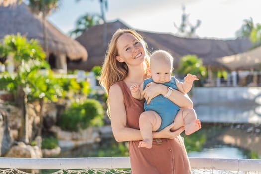 Amidst tropical palms and thatched roofs, a loving mom embraces her baby, sharing warmth and affection in a tranquil outdoor setting.