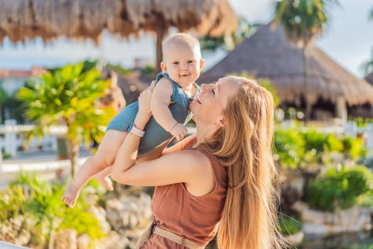 Amidst tropical palms and thatched roofs, a loving mom embraces her baby, sharing warmth and affection in a tranquil outdoor setting.