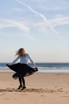 A woman is standing on the beach, wearing a blue shirt and black skirt. She is smiling and she is enjoying herself. The beach is a beautiful and peaceful setting, with the ocean in the background