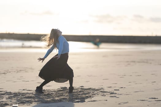 A woman is dancing on the beach in a black skirt. She is wearing a blue shirt and black boots