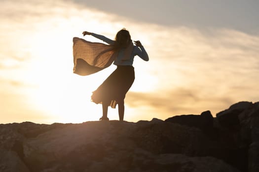 A woman is standing on a rocky hillside, with the sun setting in the background. She is wearing a scarf and she is dancing or performing a ritual. Concept of freedom and connection to nature
