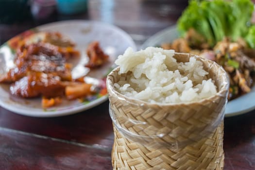 Sticky rice in sticky rice box handmade by bamboo or wicker blurred on grilled chicken and spicy minced pork salad (Laab) is a Thai carbohydrate food for health on a wood table at Thai street food
