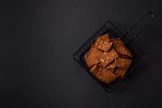 Crispy homemade brown bread chips with seeds and grains, salt, spices and herbs on a dark concrete background