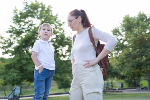 A woman and a child are standing in a park. The woman is looking at the child with a stern expression