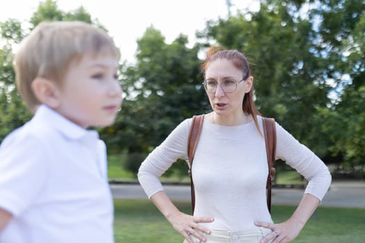 A woman is talking to a young boy in a park. The woman is wearing glasses and a backpack. Scene is tense and serious