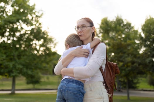 A woman is hugging a child in a park. The woman is wearing glasses and a white shirt. The scene is peaceful and heartwarming