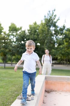 A young boy is walking on a wall in a park. A woman is watching him. The boy is wearing a white shirt and blue jeans