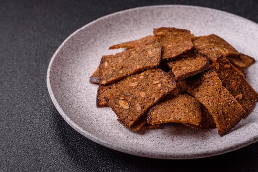 Crispy homemade brown bread chips with seeds and grains, salt, spices and herbs on a dark concrete background