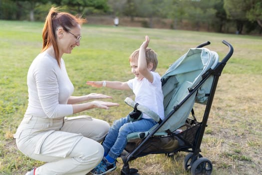 Happy boy sitting in a stroller is raising his arms in the air while playing with his smiling mother in the park