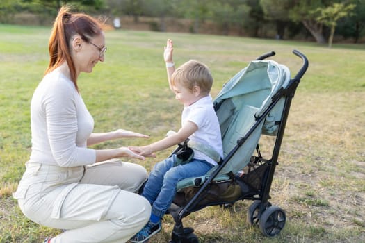 Mother is crouching and playing patty-cake with her son who is sitting in a stroller in a park