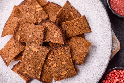 Crispy homemade brown bread chips with seeds and grains, salt, spices and herbs on a dark concrete background
