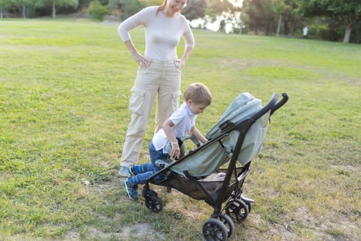 Young boy is bending over a stroller in a grassy park, while his mother stands behind him with her hands on her hips