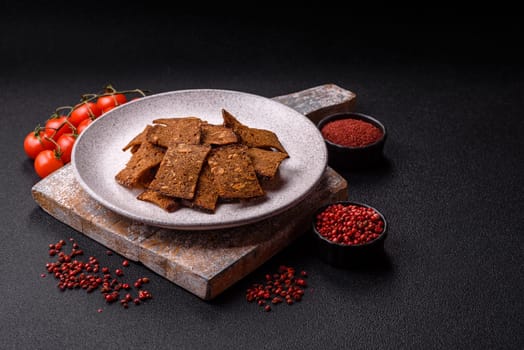 Crispy homemade brown bread chips with seeds and grains, salt, spices and herbs on a dark concrete background