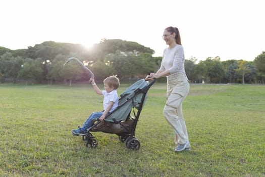 Mother is pushing her son in a stroller in a park while he is playing with a toy