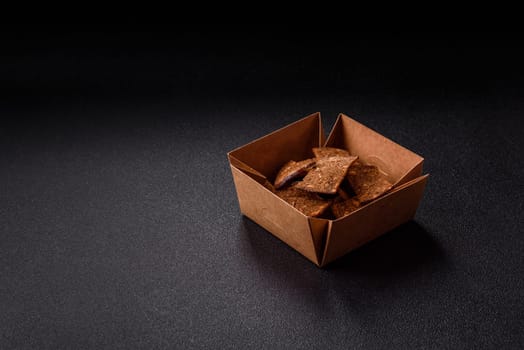 Crispy homemade brown bread chips with seeds and grains, salt, spices and herbs on a dark concrete background
