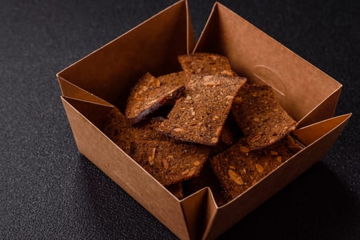 Crispy homemade brown bread chips with seeds and grains, salt, spices and herbs on a dark concrete background