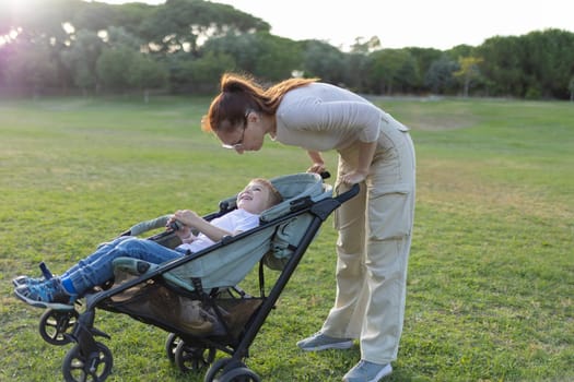 Mother is talking with her happy son who is sitting in a stroller in a park