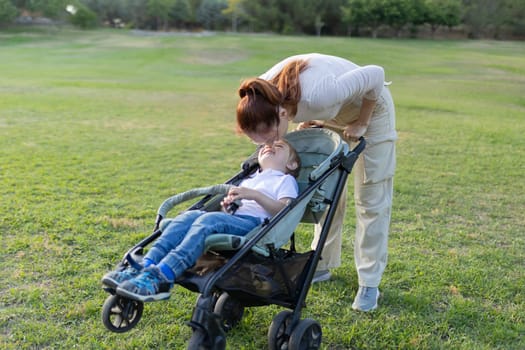Mother is sharing a sweet moment with her son who is relaxing in his stroller in a grassy park