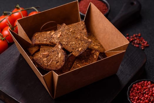 Crispy homemade brown bread chips with seeds and grains, salt, spices and herbs on a dark concrete background
