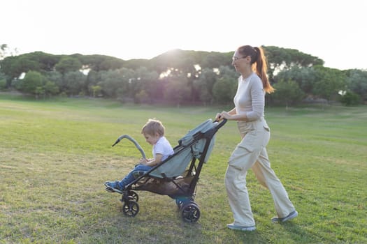 Mother is pushing her toddler son in a stroller while walking through a grassy park at sunset