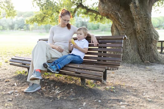 Mother and son are enjoying a peaceful moment together on a park bench, the child eating a snack