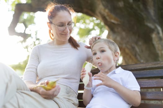 Mother smiles at her son while sitting on a bench and eating bananas
