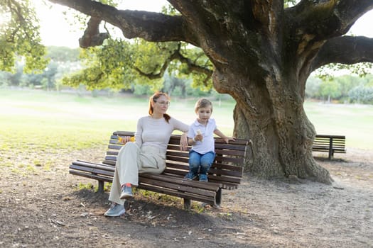 Mother and her son are enjoying each other's company while sitting on a park bench under a large tree