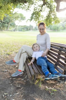 Mother sits on a park bench on a summer day while her son lays across her lap