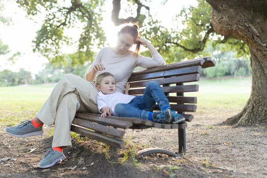 Mother is stroking her son's hair as he lays on her lap while they sit on a park bench
