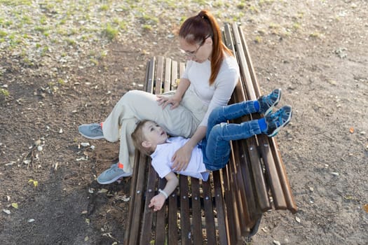 Mother is sitting on a bench in the park, talking to her young son who is lying down on the bench