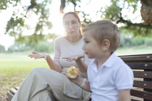 Mother is gesturing with her hands while talking to her son, who is holding a snack, in a park