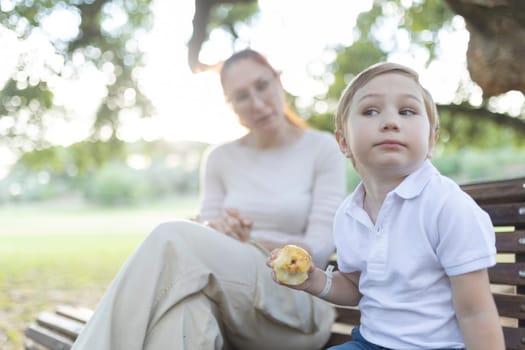 Thoughtful little boy holding a half-eaten apple sitting on a bench in the park with his mom, enjoying a sunny day outdoors