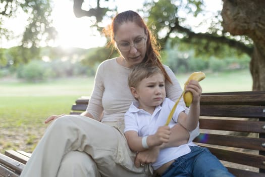 Young child is holding a banana while talking with his mother on a park bench in the summer
