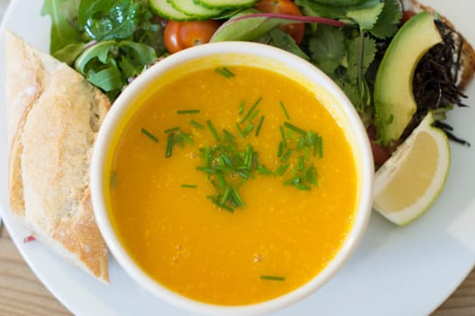 Yellow pumpkin soup with fresh herbs in a bowl, top view on a plate with fresh vegetable salad and pieces of white bread,
