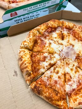 Castle Rock, Colorado, USA-April 8, 2023-A freshly baked cheese pizza with golden crust and melted mozzarella cheese sitting in an open delivery box The pizza is pre sliced and includes a pizza saver in the center In the background another pizza box is visible showing branding and ordering information The setting appears to be a casual dining or takeout environment perfect for a quick meal or gathering
