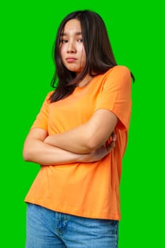 A young woman stands confidently with her arms crossed, wearing an orange shirt and blue jeans. Her serious expression contrasts with the vibrant green backdrop, drawing attention.
