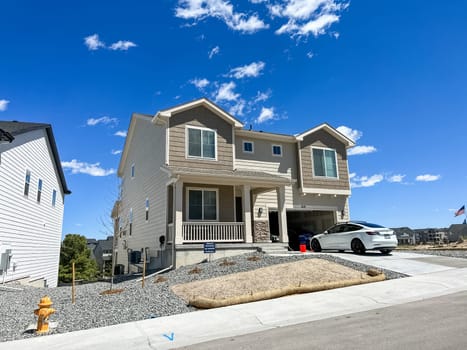 Castle Rock, Colorado, USA-May 6, 2023-A modern two story suburban home with a beige and white exterior featuring a front porch and an attached garage A white car is parked in the driveway while the front yard is landscaped with gravel and a patch of dried grass The sky is clear blue with scattered clouds creating a bright and inviting atmosphere