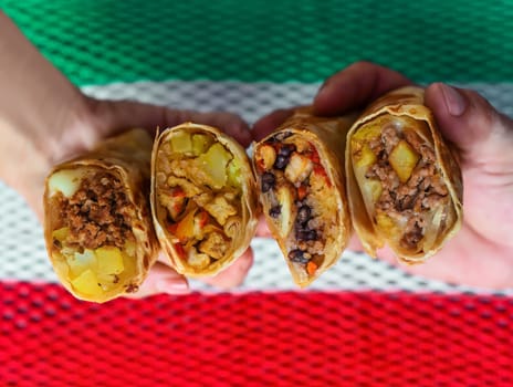 Hands holding Mexican burritos with chorizo, beef, chicken, vegetarian against mexican flag