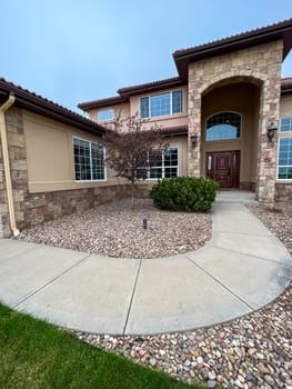 Castle Pines, Colorado, USA-May 10, 2023-A stunning luxury home featuring a grand entrance with an arched entryway stone exterior and large windows The front yard is landscaped with decorative rocks bushes and a curved concrete walkway leading to the front door The house has a Mediterranean-style roof and elegant outdoor lighting fixtures