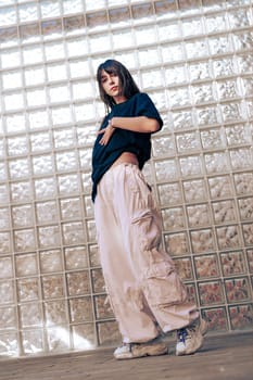 A young woman showcases her dance skills in a stylish outfit featuring loose pants and a t-shirt. She poses confidently in front of a textured glass wall in a bright setting.