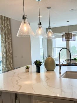 Castle Rock, Colorado, USA-May 14, 2023-A beautifully designed modern kitchen island featuring a white marble countertop with elegant veining Three glass pendant lights hang above the island providing warm illumination The space is decorated with small potted plants a stylish vase and a sleek stainless steel faucet Large windows in the background let in natural light enhancing the open and airy feel of the kitchen