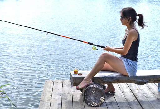 Young fisherwoman sitting on wooden pier, fishing in the lake.