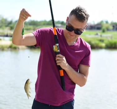 Young fisherman holding crucian carp on fishing rod against a pond