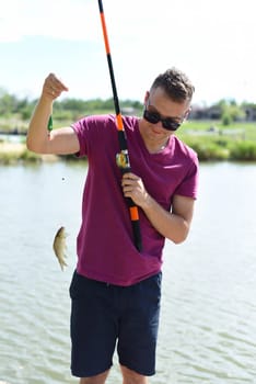 Young fisherman holding crucian carp on fishing rod against a pond