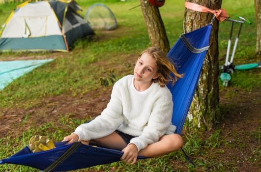 Little blonde curly girl sitting in hammock in the camping trip in forest in autumn